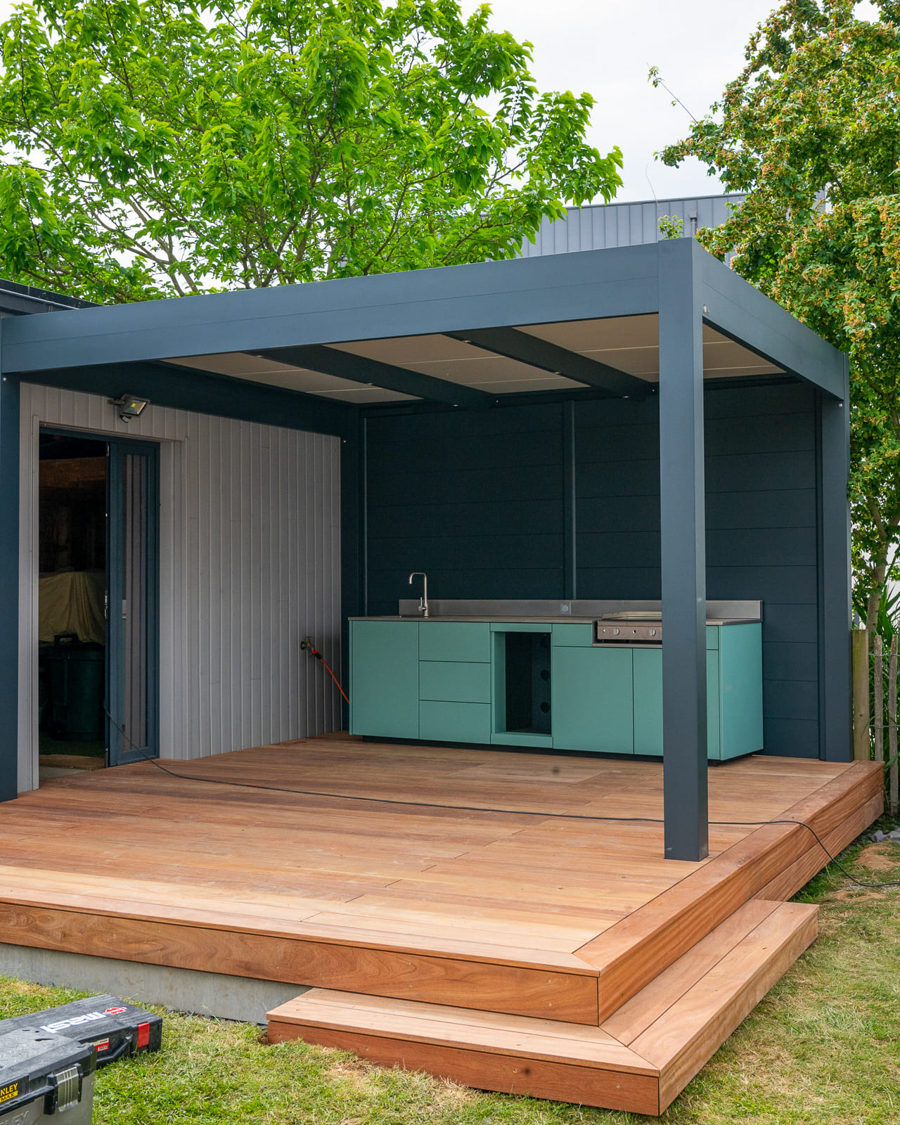 Cuisine extérieure bleue avec plancha et réfrigérateur intégrés, sous pergola sur terrasse bois Cuisine extérieure bleue avec plancha et réfrigérateur intégrés, sous pergola sur terrasse bois
